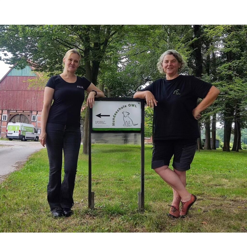 Julia Becker und Ingrid Schnusenberg posieren vor dem Hundeschule-OWL Schild an lässig des Führungswechsels von Ingrid an Julia zum Oktober 2024